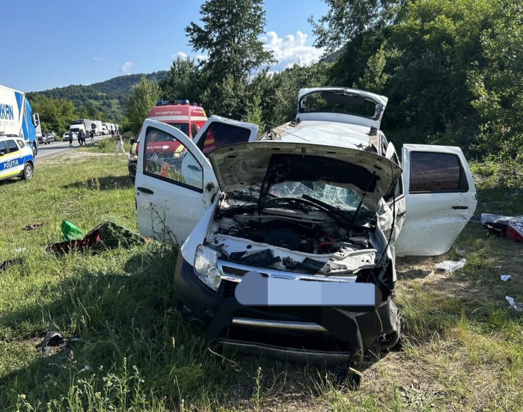 White SUV with front-end damage and open hood on a grassy roadside; emergency responders and police vehicles in background.