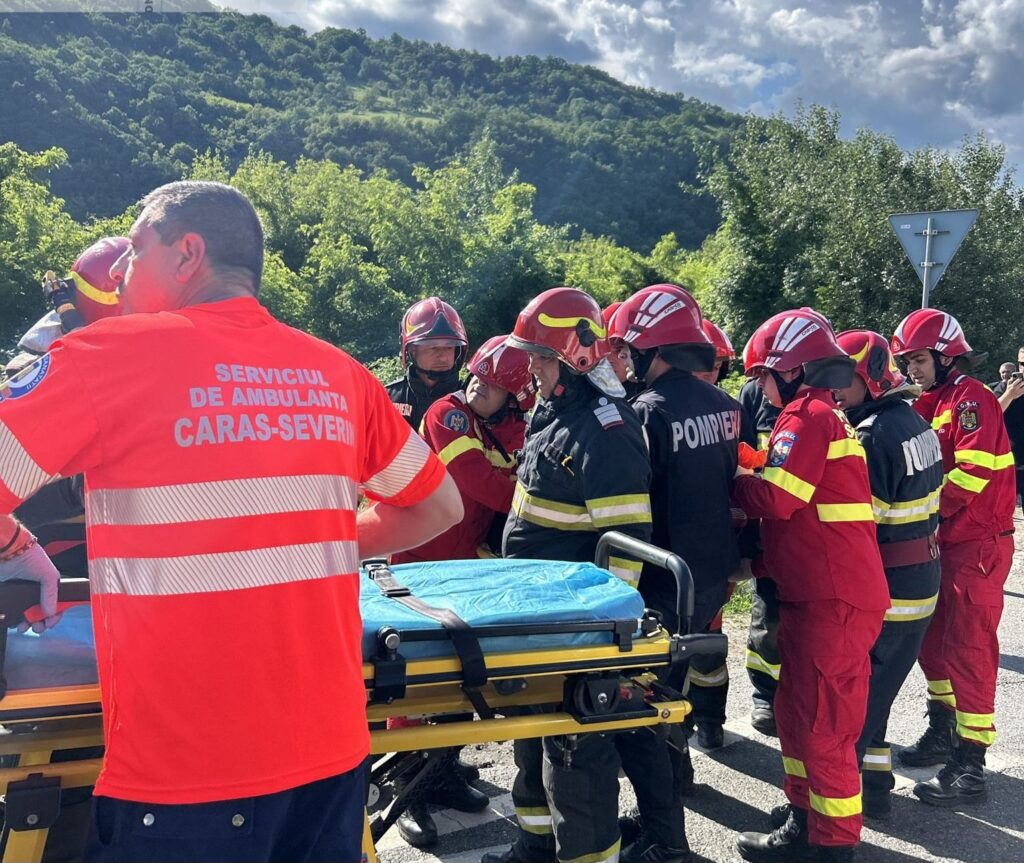 Group of firefighters in red and black uniforms cluster around a wheeled stretcher on a road, preparing to transfer a patient outside.