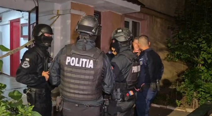 Nighttime scene of Romanian police in tactical gear surrounding and restraining a man outside a residential building.