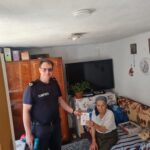 Firefighter in a dark blue uniform stands beside an elderly woman seated on a bed, handing her a document in a cozy living room.