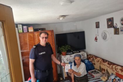 Firefighter in a dark blue uniform stands beside an elderly woman seated on a bed, handing her a document in a cozy living room.