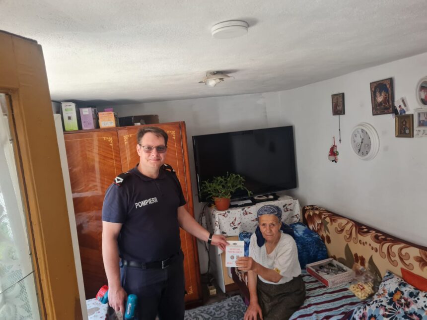 Firefighter in a dark blue uniform stands beside an elderly woman seated on a bed, handing her a document in a cozy living room.
