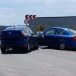 Two blue cars have collided head-on on a rural road, debris scattered on the pavement, with a 30 mph sign and a distant building in view.