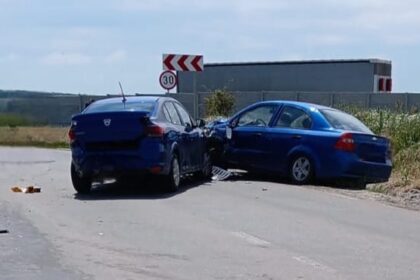 Two blue cars have collided head-on on a rural road, debris scattered on the pavement, with a 30 mph sign and a distant building in view.