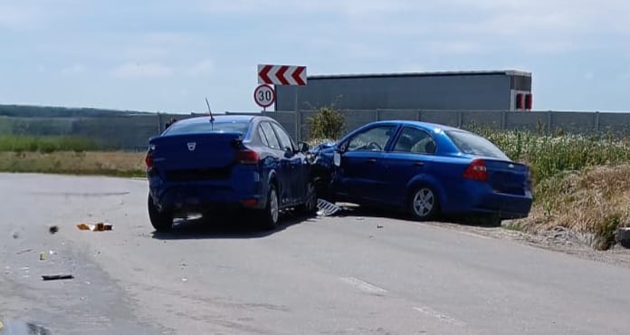 Two blue cars have collided head-on on a rural road, debris scattered on the pavement, with a 30 mph sign and a distant building in view.