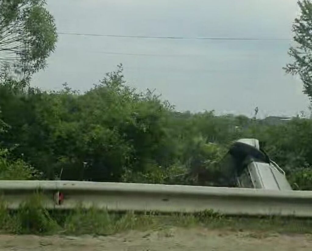 Silver sedan crashed into a guardrail along a grassy roadside, with dense bushes in the background.