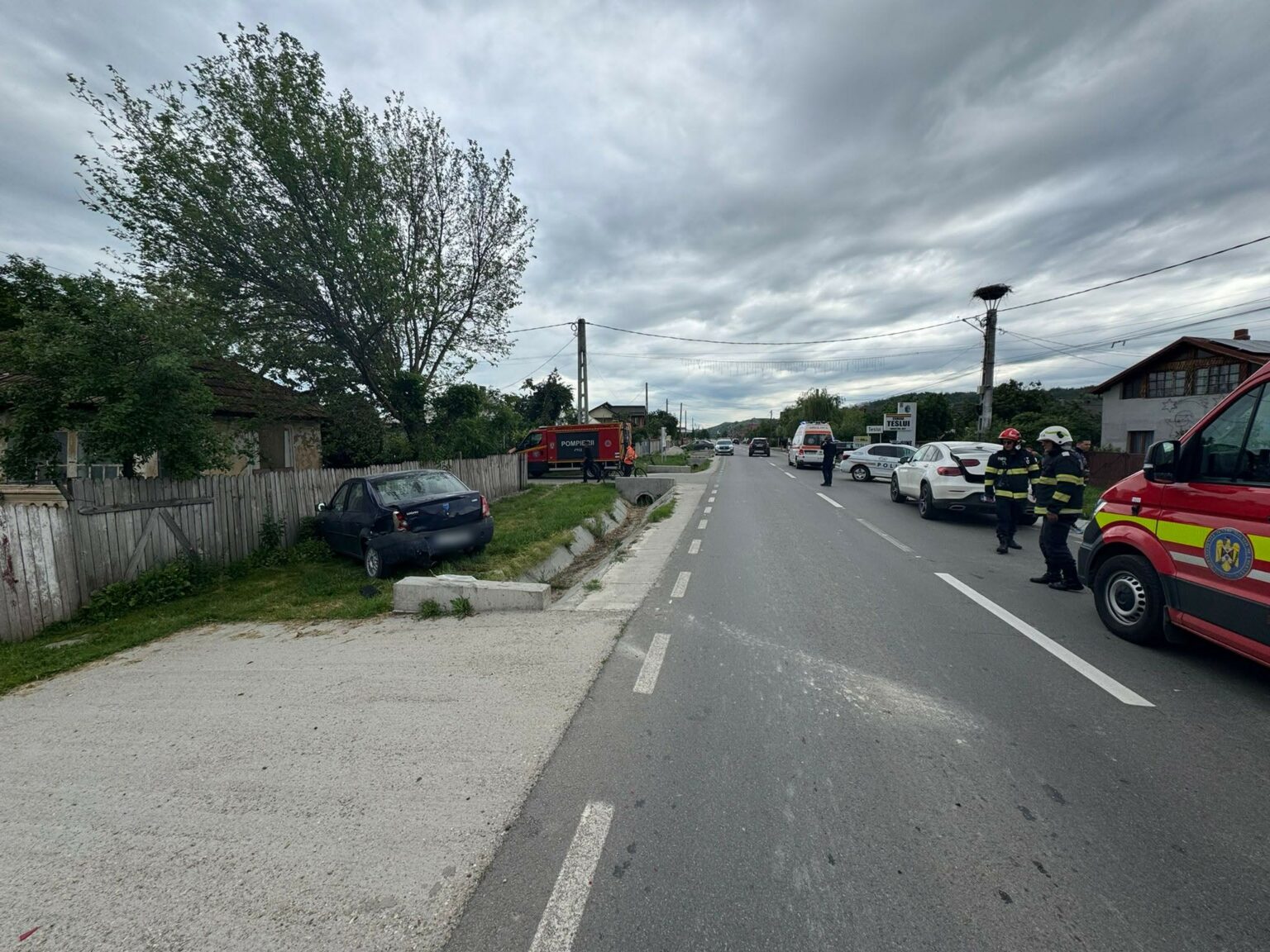 Emergency responders, including firefighters and police, gather on a two-lane street near a damaged dark car after a crash, with a red fire truck in the background and an overcast sky above the scene.