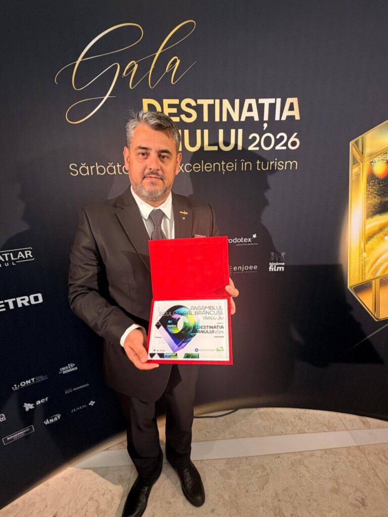 Man in a dark suit holding a red award box and certificate, posing in front of a banner at a formal event.