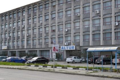 Large gray office building with many windows and wall-mounted air conditioners, a parking lot in front, and a blue-and-white bus shelter on the right.