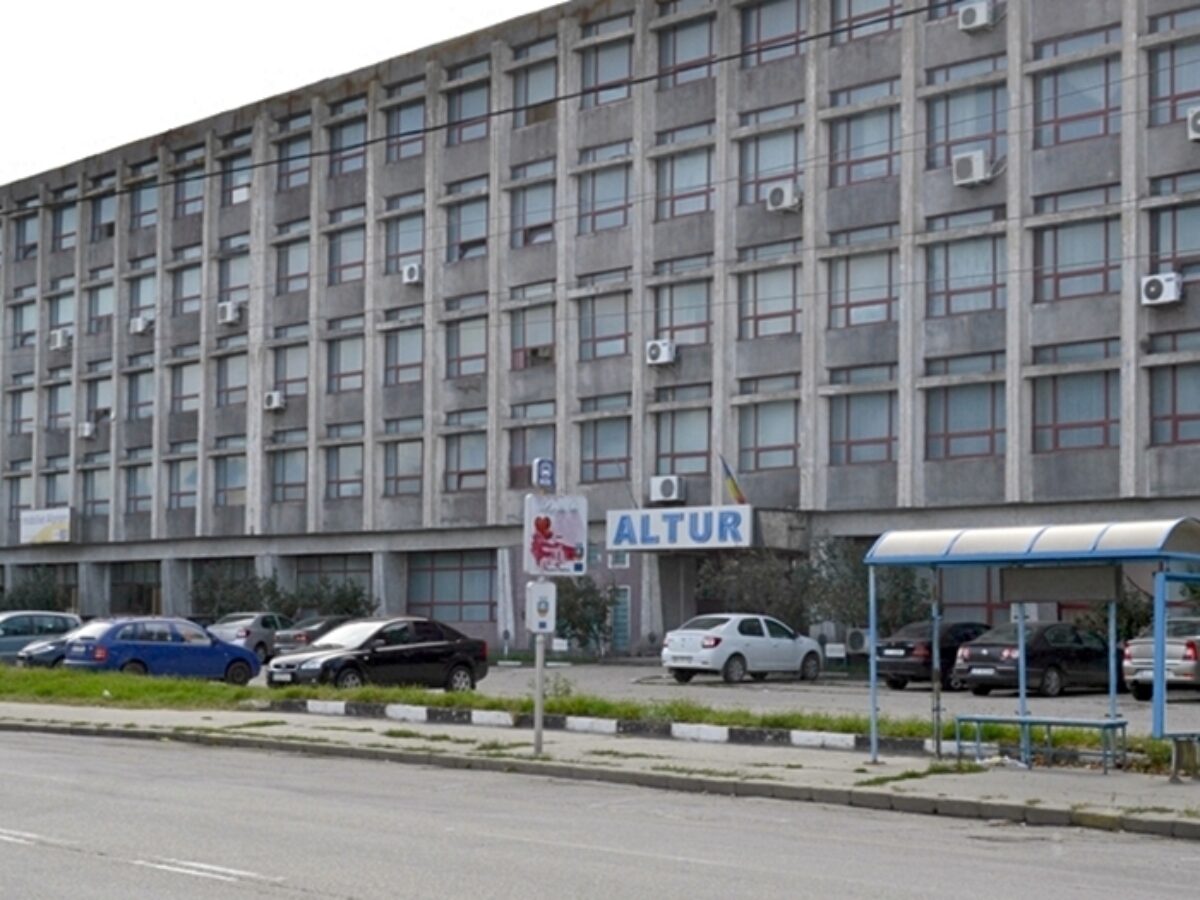 altur_slatina-1200x900 - Jurnalul Olteniei – Știri din Craiova și Oltenia Large gray office building with many windows and wall-mounted air conditioners, a parking lot in front, and a blue-and-white bus shelter on the right.