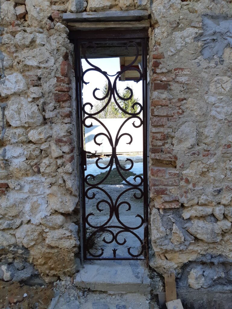 Decorative black wrought-iron gate with scrollwork set in a crumbling stone wall, opening to a snowy outdoor scene beyond.