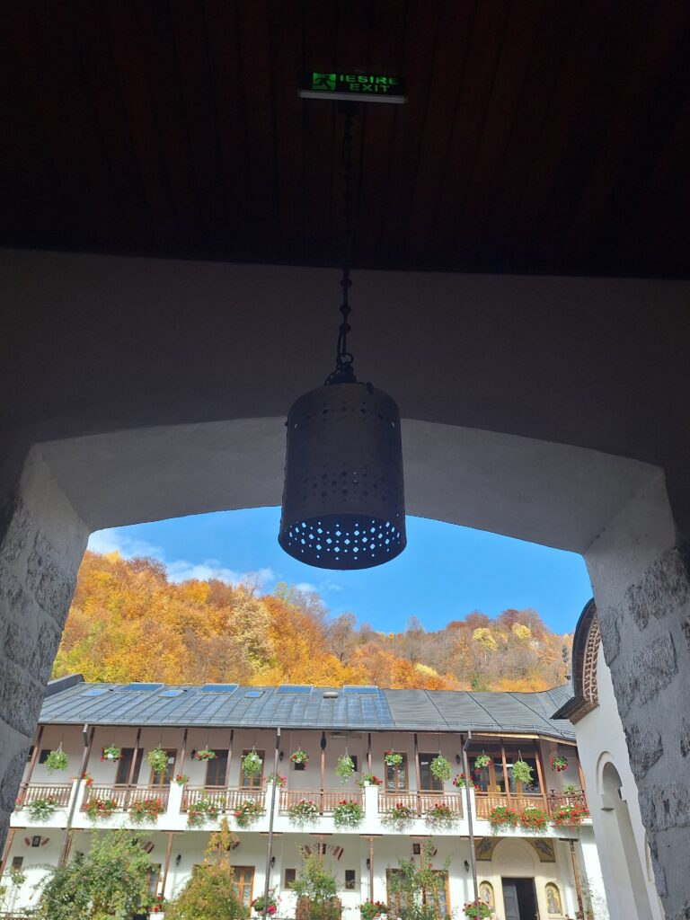 Hanging perforated metal lantern suspended from a dark ceiling over a stone archway, opening to a sunny courtyard with flowering balconies and autumn trees outside.