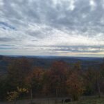 Hilly countryside with autumn trees under a cloudy sky; a fence runs along the foreground.