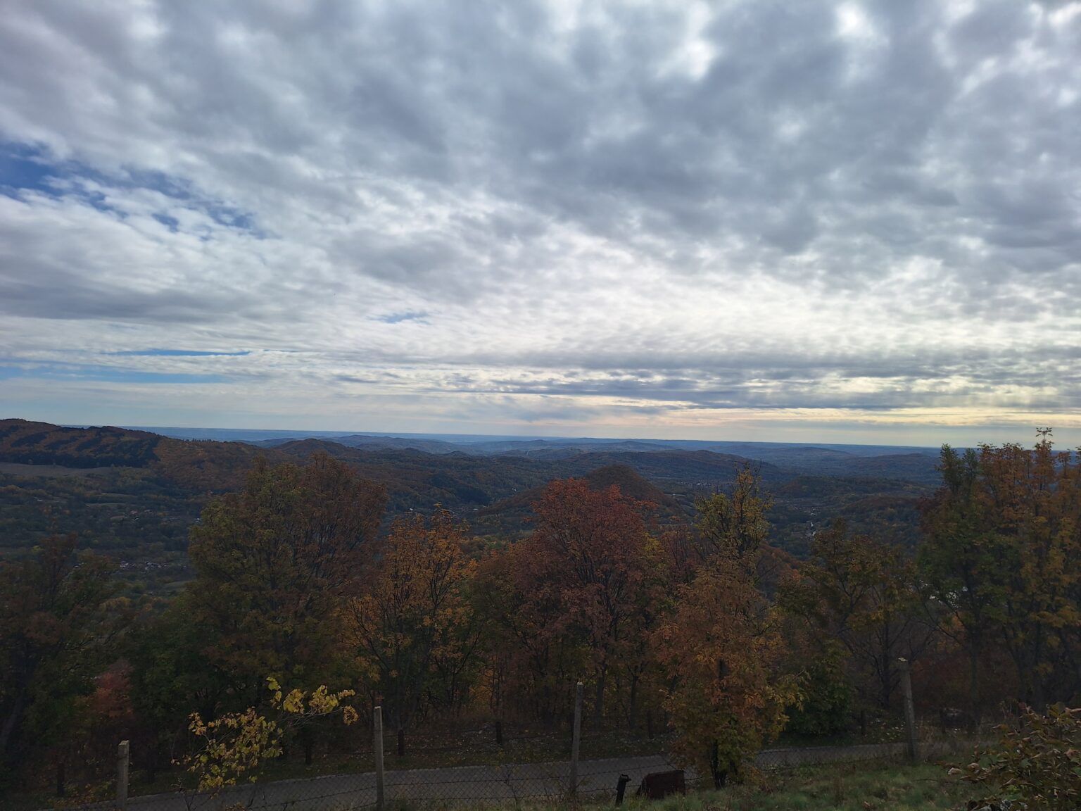 Hilly countryside with autumn trees under a cloudy sky; a fence runs along the foreground.