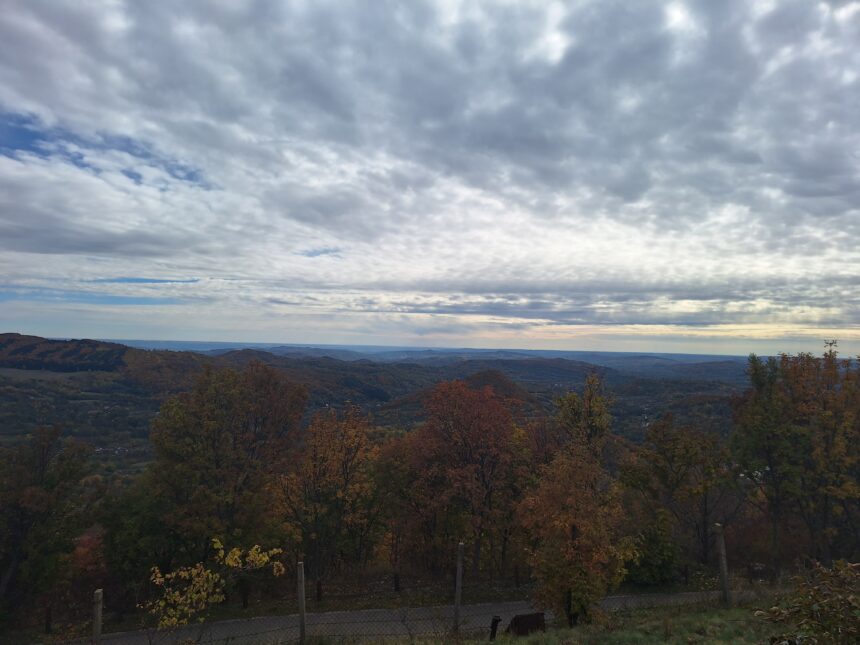 Hilly countryside with autumn trees under a cloudy sky; a fence runs along the foreground.