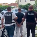 Two police officers in dark vests escort a handcuffed man down a residential sidewalk. A car is parked nearby as they walk away from the camera.