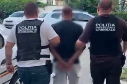 Two police officers in dark vests escort a handcuffed man down a residential sidewalk. A car is parked nearby as they walk away from the camera.