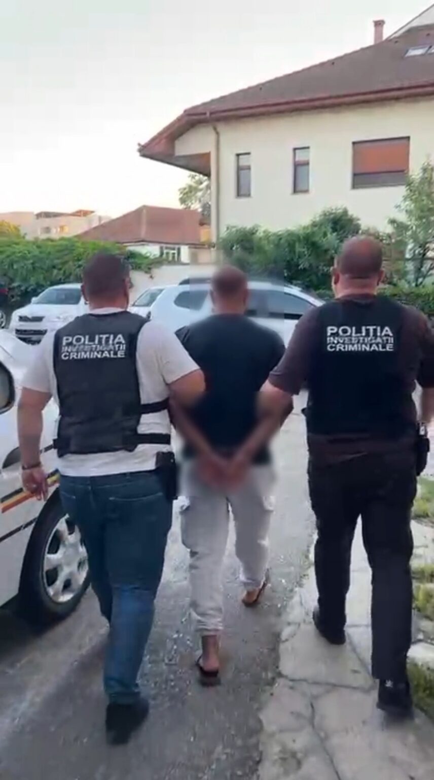 Two police officers in dark vests escort a handcuffed man down a residential sidewalk. A car is parked nearby as they walk away from the camera.