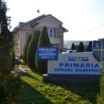 Sign for Baldovinești Commune Local Council in Olt County in front of a beige municipal building with evergreen trees nearby.