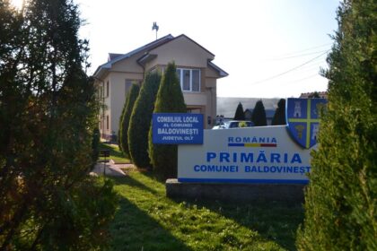 Sign for Baldovinești Commune Local Council in Olt County in front of a beige municipal building with evergreen trees nearby.