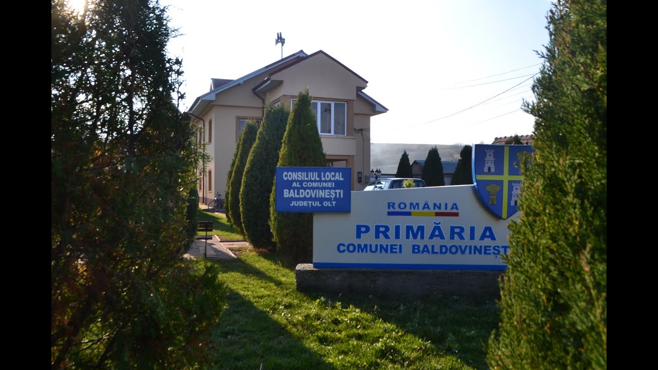 Sign for Baldovinești Commune Local Council in Olt County in front of a beige municipal building with evergreen trees nearby.