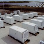 Industrial solar farm with many white shipping containers on concrete pads, fencing, and solar panels in the background