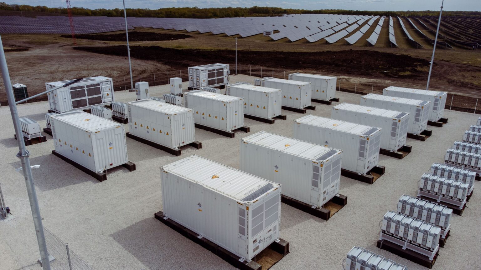 Industrial solar farm with many white shipping containers on concrete pads, fencing, and solar panels in the background