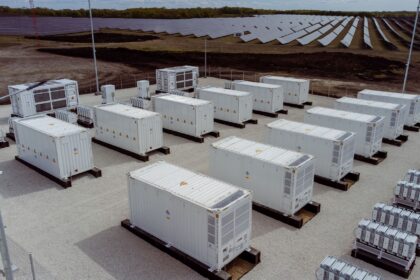 Industrial solar farm with many white shipping containers on concrete pads, fencing, and solar panels in the background