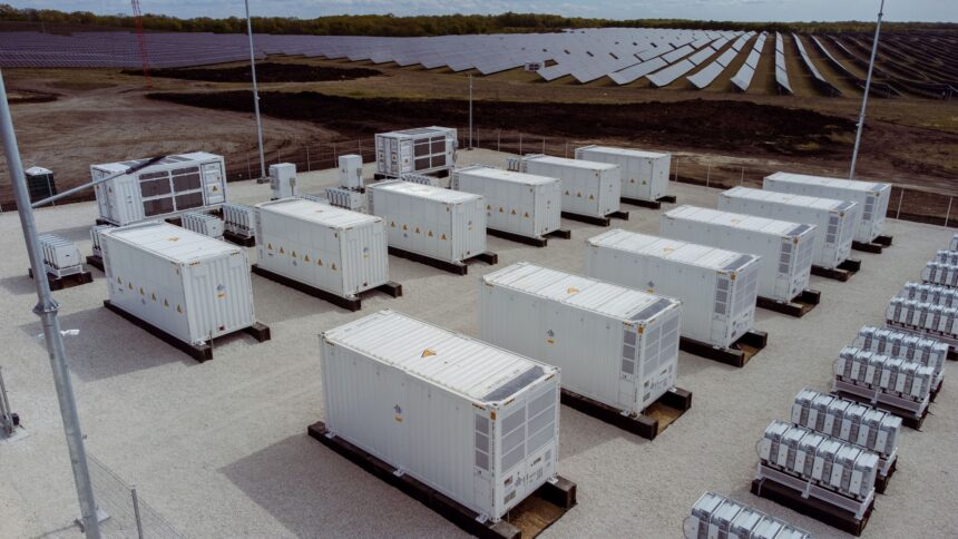Industrial solar farm with many white shipping containers on concrete pads, fencing, and solar panels in the background