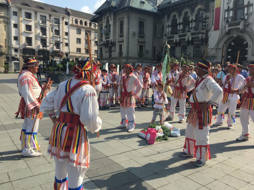 Group of people in white traditional outfits with colorful sashes and hats marching in a city square behind historic buildings.