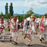 Group of performers in traditional white and red embroidered outfits dancing with wooden sticks at an outdoor festival, ribbons trailing from their costumes as spectators watch.