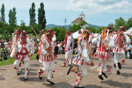 Group of performers in traditional white and red embroidered outfits dancing with wooden sticks at an outdoor festival, ribbons trailing from their costumes as spectators watch.