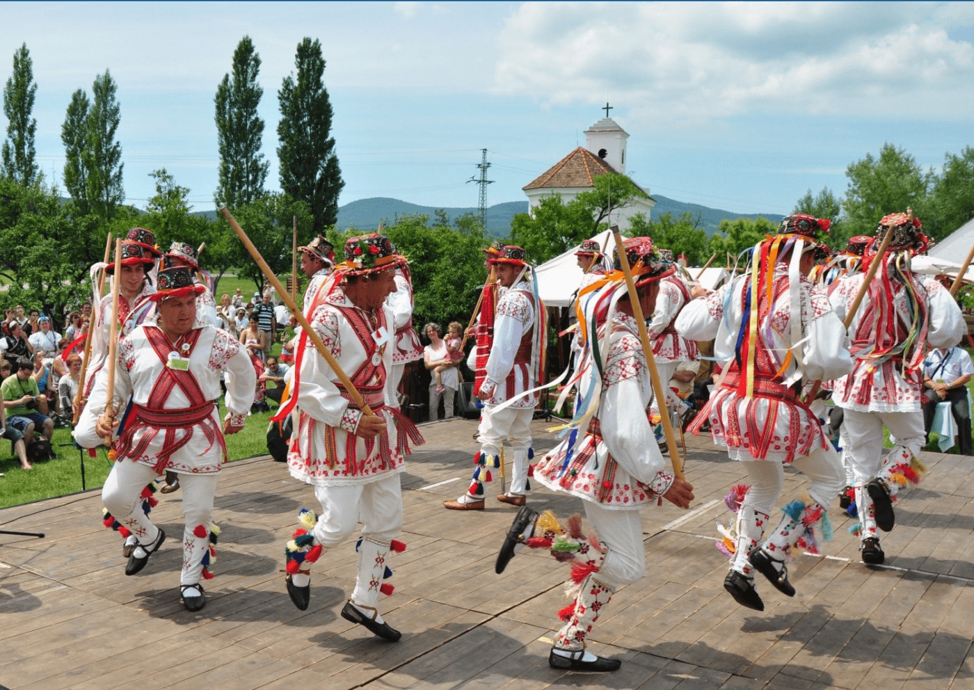 Group of performers in traditional white and red embroidered outfits dancing with wooden sticks at an outdoor festival, ribbons trailing from their costumes as spectators watch.