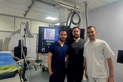 Three medical professionals in scrubs and a white coat stand in an operating room beside a large monitor displaying medical images.