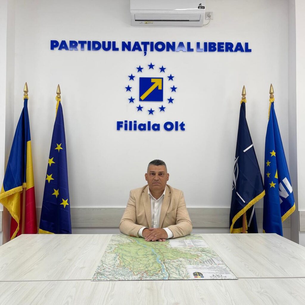 Man seated at a conference desk in a political party office, with flags on either side and a logo reading 'PARTIDUL NAȚIONAL LIBERAL' and 'Filiala Olt' on the wall behind him