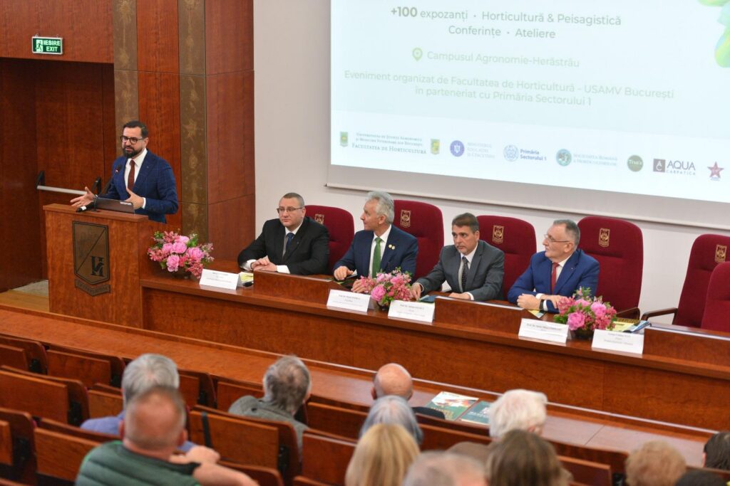 Man in a blue suit speaks at a wooden podium in a conference hall; four panelists sit at a long desk behind him with pink flowers in front of them.