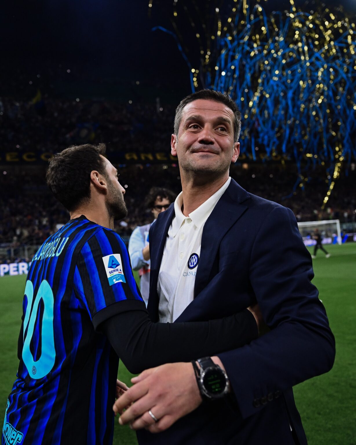 Two men embrace on a football pitch during celebration, one in a dark suit and the other in a blue striped jersey with a crowd and confetti in the background.