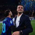 Two men embrace on a football pitch during celebration, one in a dark suit and the other in a blue striped jersey with a crowd and confetti in the background.