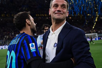 Two men embrace on a football pitch during celebration, one in a dark suit and the other in a blue striped jersey with a crowd and confetti in the background.