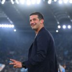Sports coach in a navy blazer smiling on a brightly lit soccer stadium field with a cheering crowd in the background.