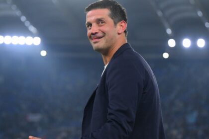 Sports coach in a navy blazer smiling on a brightly lit soccer stadium field with a cheering crowd in the background.