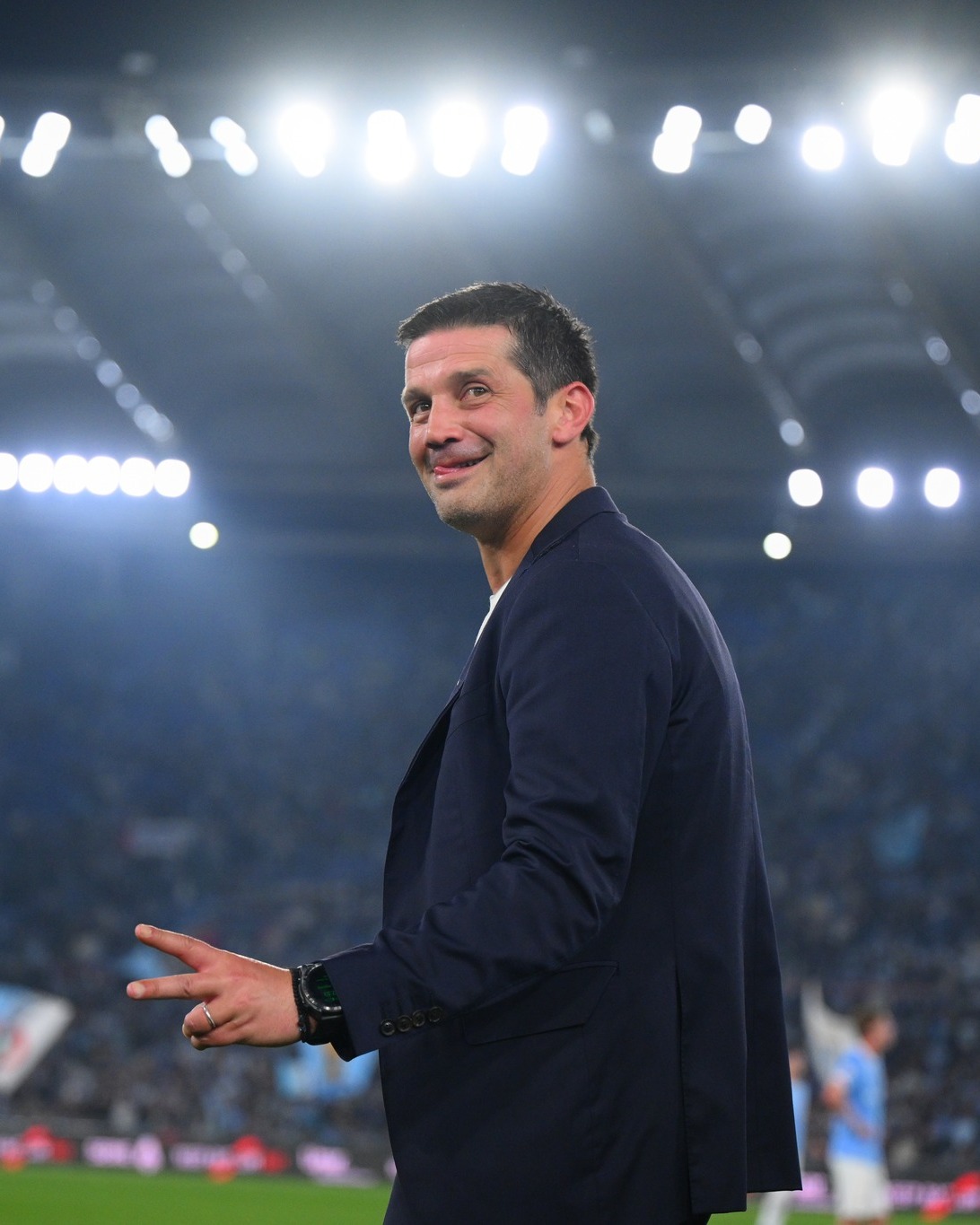 Sports coach in a navy blazer smiling on a brightly lit soccer stadium field with a cheering crowd in the background.