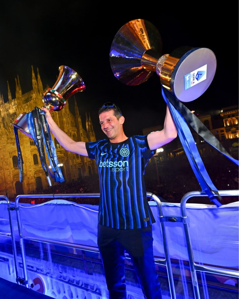 Man wearing a blue striped jersey raises two large trophies with ribbons at night, celebrating victory in a stadium.