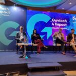 Panel of four speakers seated on a stage during Govtech 4 Impact World Congress, with a blue, illuminated backdrop and large event branding behind them.