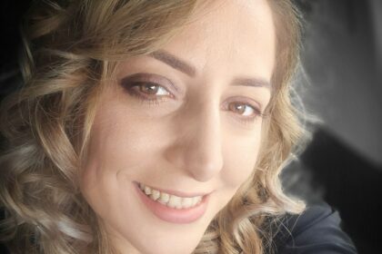 Close-up portrait of a smiling woman with light brown, wavy hair, inside a car wearing a dark top.