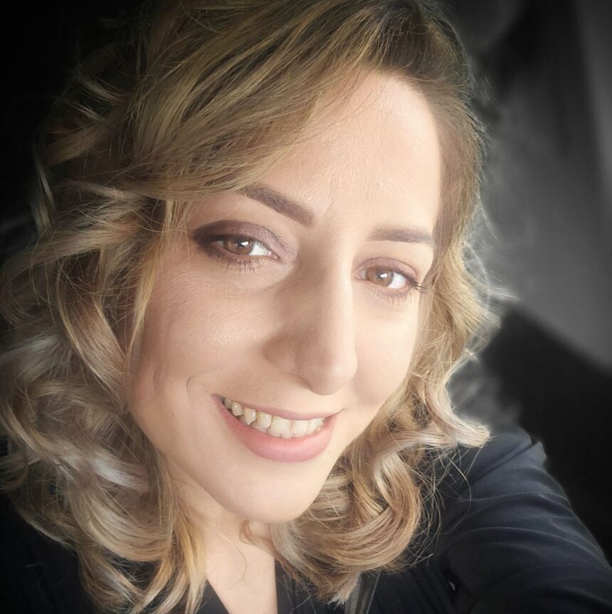 Close-up portrait of a smiling woman with light brown, wavy hair, inside a car wearing a dark top.