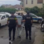 Two police officers in dark uniforms escort a person down a wet street; police cars parked nearby (street scene).