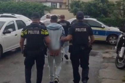 Two police officers in dark uniforms escort a person down a wet street; police cars parked nearby (street scene).