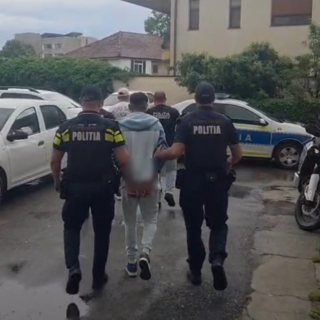 Two police officers in dark uniforms escort a person down a wet street; police cars parked nearby (street scene).