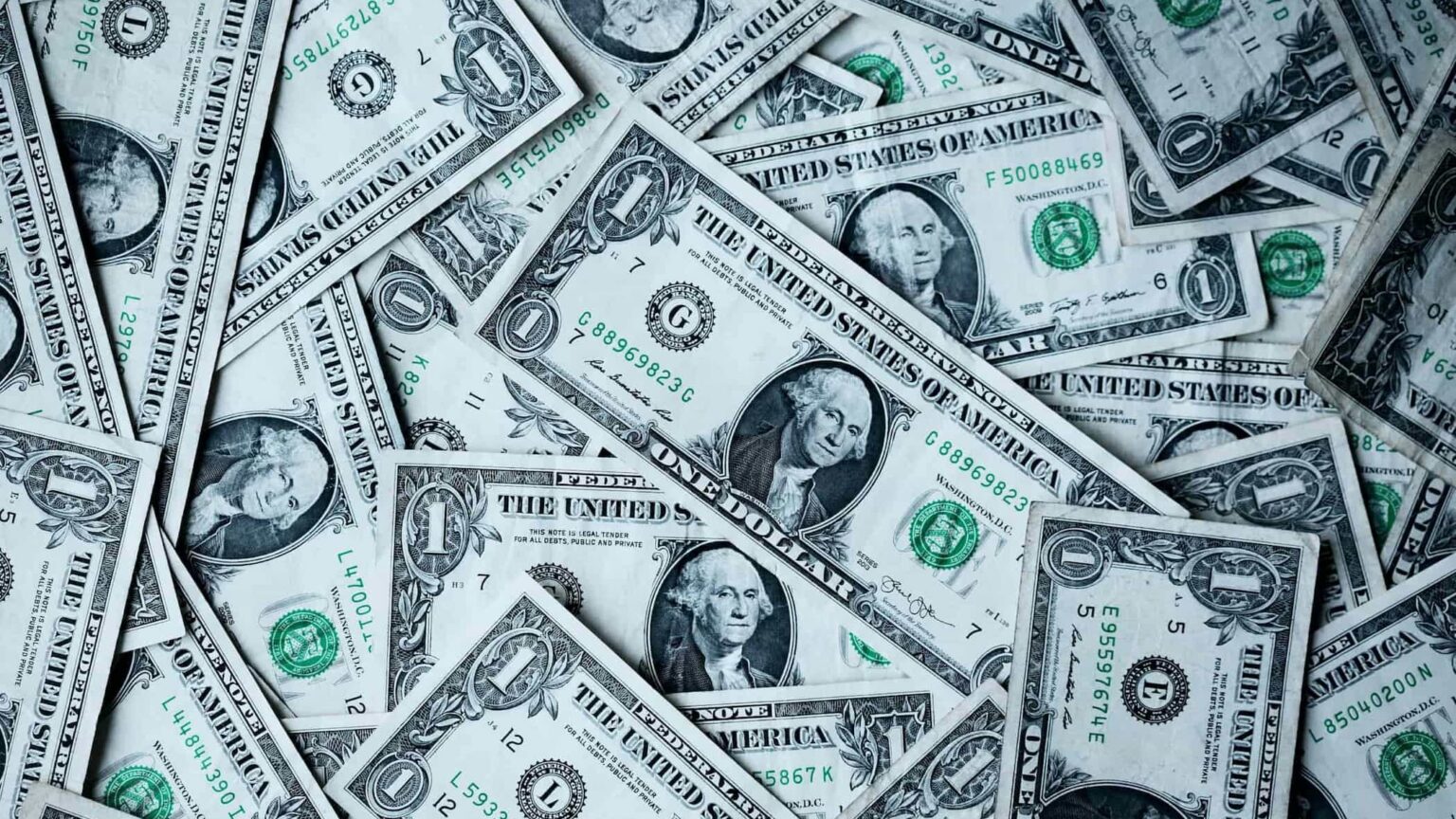 Pile of scattered one-dollar bills showing portraits of George Washington and green seals and serial numbers in a money-filled scene.